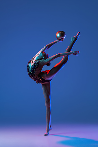 Perfect body flexibility. One young girl, rhythmic gymnastics artist in bright beautiful sports costume isolated on blue background in neon. Concept of sport, action, aspiration, active lifestyle.