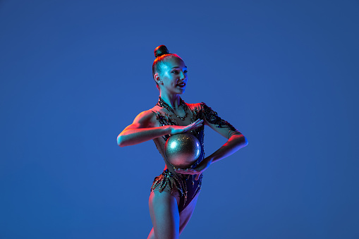 Beginning of performance. One young girl, rhythmic gymnastics artist in bright beautiful sports costume isolated on blue studio background in neon. Concept of sport, action, aspiration, active lifestyle.