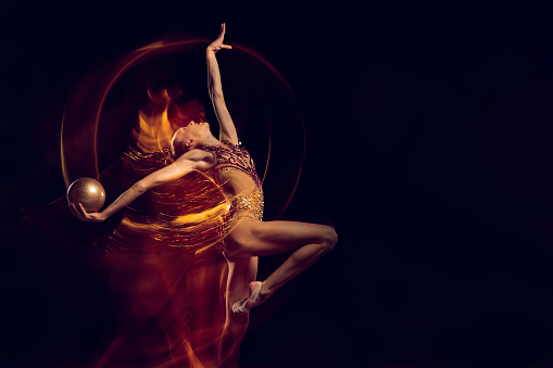 Dancing with golden ball. Young girl, female rhythmic gymnastics artist in action isolated on dark studio background with mixed light. Concept of sport, action, aspiration, beauty and achievements.