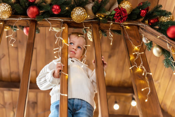 portrait cute little blond toddler boy stand looking at house wooden staricase handrails railings decorated with christmas tree and golden lights garland. xmas family celebration christmas concept - christmas child little boys peeking imagens e fotografias de stock