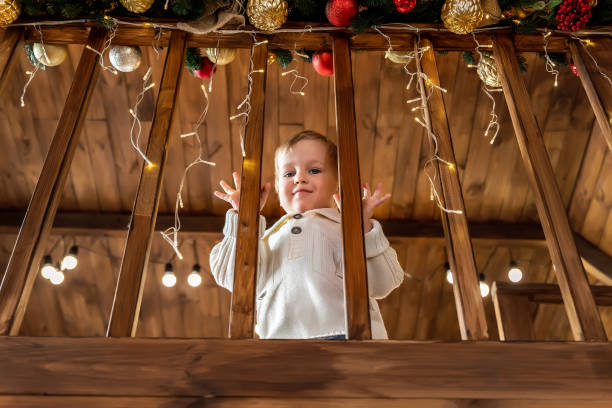portrait cute little blond toddler boy stand looking at house wooden staricase handrails railings decorated with christmas tree and golden lights garland. xmas family celebration christmas concept - christmas child little boys peeking imagens e fotografias de stock