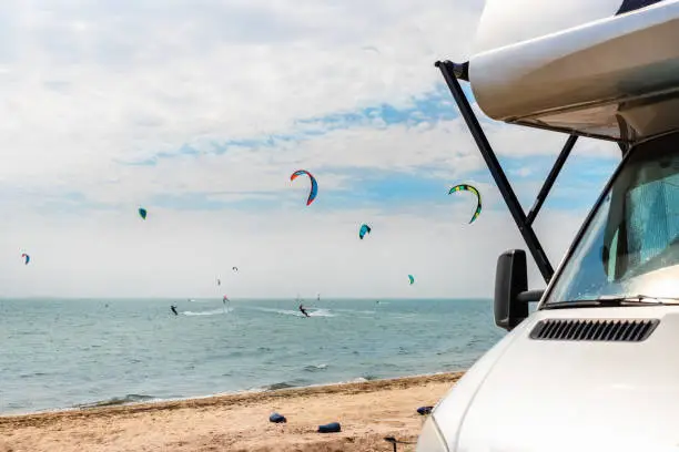 Panoramic view of many surf board kite riders on sand beach watersport spot on bright sunny day against rv camper van vehicle at sea ocean coast at surfing camp. Fun adventure travel sport acitivity.