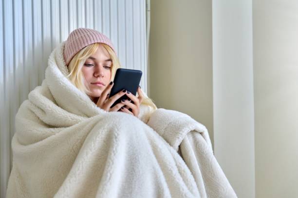 Cold season, frozen teenager in hat under blanket sitting near a heating radiator with smartphone stock photo