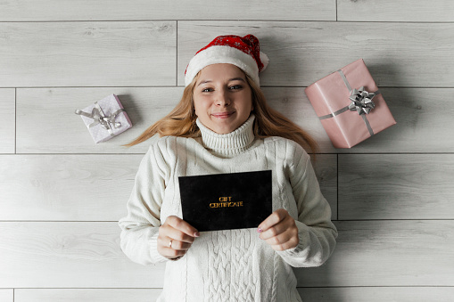 A woman in a white sweater lies on the floor and holds a gift certificate in her hands. High quality photo