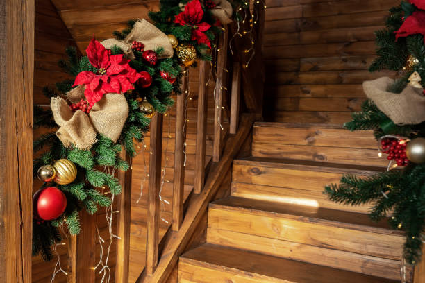 Detail house wooden staricase handrails railings decorated with artificial holly poinsettia flower, burlap bow, christmas tree and golden lights garland. Xmas family home interior decor idea concept. detalle casa de madera staricase barandillas decoradas con flor de pascua de acebo artificial, arco de arpillera, árbol de navidad y guirnalda de luces doradas. concepto de idea de decoración de interiores para el hogar familiar de navidad