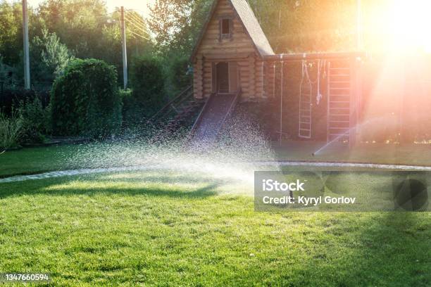 Landscape Automatic Garden Watering System With Different Sprinklers Installed Under Turf Landscape Design With Lawn Hills And Fruit Garden Irrigated With Smart Autonomous Sprayers At Sunset Time Stock Photo - Download Image Now
