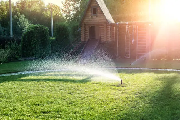 Landscape automatic garden watering system with different sprinklers installed under turf. Landscape design with lawn hills and fruit garden irrigated with smart autonomous sprayers at sunset time.