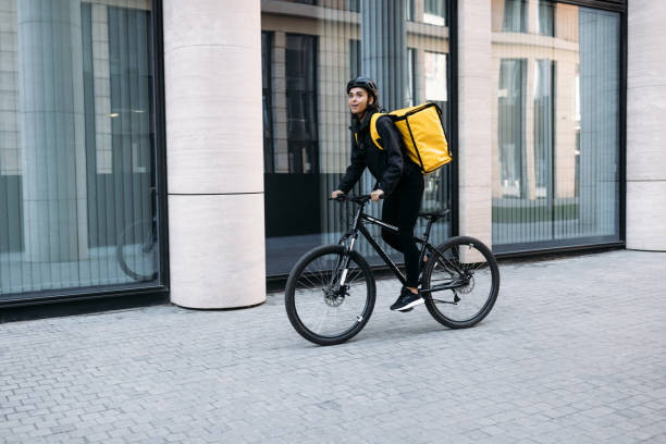istock Smiling courier girl riding a bicycle wearing delivery thermal backpack looking for a customer address 1347664737