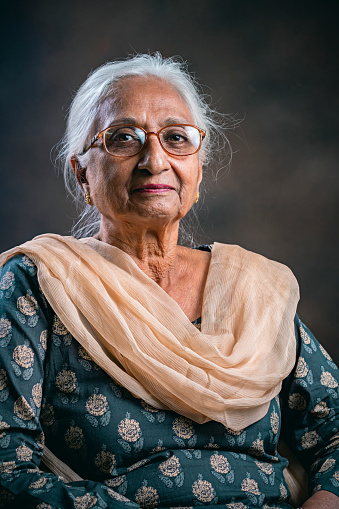 In this indoor studio portrait, an Asian/Indian real senior woman sits in a studio against the smoky dark background and looks at the camera with a blank expression.