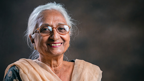 In this indoor studio portrait, an Asian/Indian real senior woman sits in a studio against the smoky dark background and looks at the camera with a toothy smile.