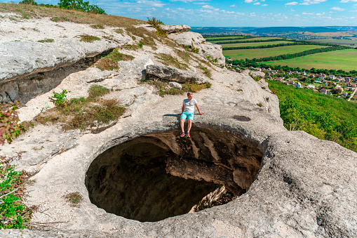 A young man on the edge of a precipice in the form of a huge hole in the ground among green fields and mountains on a sunny summer day. An abnormal cave, a sinkhole in the ground.