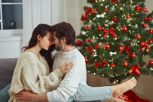Marry Christmas Holidays and Happy New Year. Couple in love embracing on the sofa near the Christmas tree. Romantic tender couple in sweaters celebrating New Year at home near decorated fir tree and various presents.