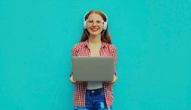 Portrait of modern young woman working with laptop listening to music in headphones on blue background stock photo