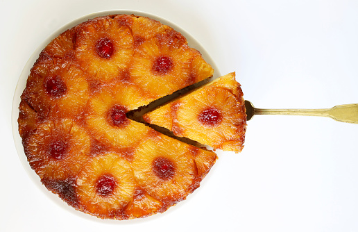 Cutted pineapple upside down cake with glacé cherry and scapula on white background. Directly above.