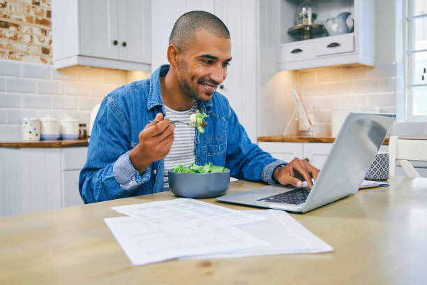 istock Shot of a young man using a laptop and having a healthy salad while working at home 1347684119