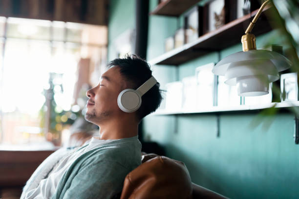 young asian man with eyes closed, enjoying music over headphones while relaxing on the sofa at home - spiritualiteit stockfoto's en -beelden