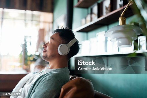 istock Young Asian man with eyes closed, enjoying music over headphones while relaxing on the sofa at home 1347685599