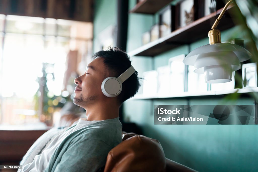 Young Asian man with eyes closed, enjoying music over headphones while relaxing on the sofa at home Relaxation Stock Photo