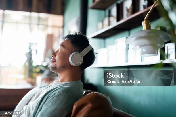 Young Asian Man With Eyes Closed Enjoying Music Over Headphones While Relaxing On The Sofa At Home Stock Photo - Download Image Now