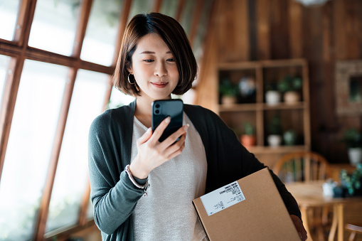 Smiling young Asian woman shopping online with smartphone on hand, receiving a delivered parcel by home delivery service. Online shopping, online banking. Enjoyable customer shopping experience