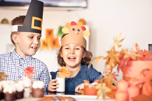 Kids sitting on festive table and celebrating Thanksgiving day. Children in paper turkey hat and pilgrim hats eating cupcakes and drinking milk. stock photo