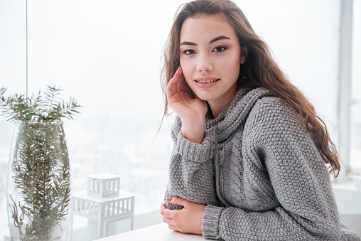 Picture of cheerful young woman dressed in sweater sitting in cafe at cold winter day. Look at camera.