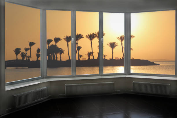 Panoramic window overlooking dawn above tropical island in ocean. Sunset above sea. Travel concept. Summer vacations. Tropical resort. Wide window with seascape. Palm trees on island. View from room