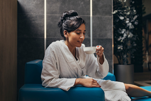 Smiling female in bathrobe drinking a cup of tea while sitting on a blue sofa