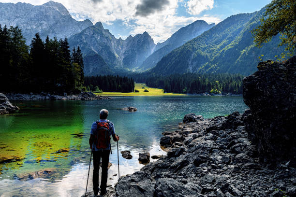 An elderly man walks around Lake Fusine with high rocky Mangart mountains in the background, Julian Alps, Italy Magical mountain landscape and touristic place. Admirable superior Fusine lake and Mangart mountain in background, Julian Alps, Tarvisio, Udine region, Friuli Venezia Giulia, Italy, Europe julian alps stock pictures, royalty-free photos & images