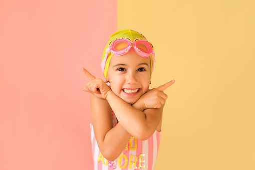 Cute adorable funny girl in a trendy swimming glasses and a pool cap, pointing fingers in different sides at copy space on an pink and yellow studio background. Child indicating at discount or offer