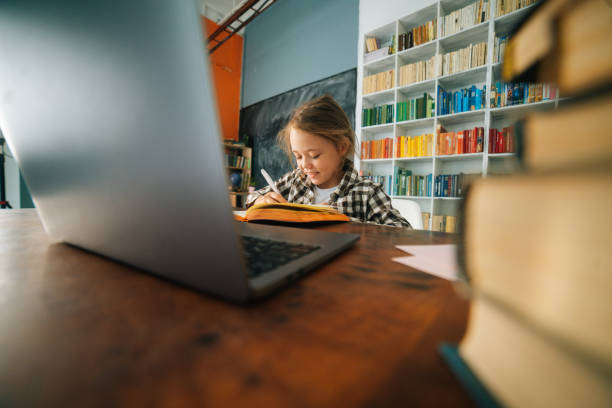 istock Smiling positive primary child school girl doing homework writing notes in paper notebook sitting at table with laptop. 1347714871