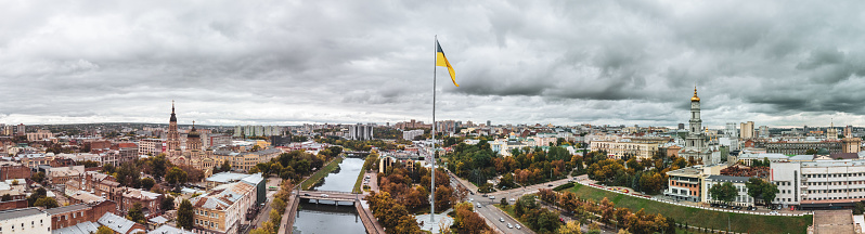 Flag of Ukraine on flagpole with epic heavy cloudscape, wide city aerial panorama on autumn river Lopan embankment, Holy Annunciation Cathedral, Dormition Cathedral in Kharkiv, Ukraine