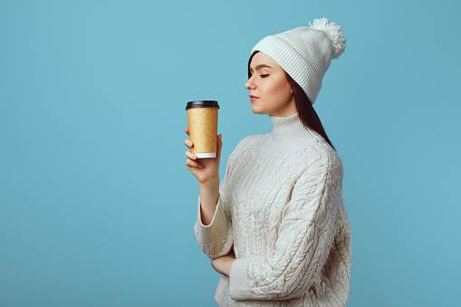 Young girl wearing white hat and sweater, enjoys drinking coffee from takeout cup, has cheerful expression, poses over blue background