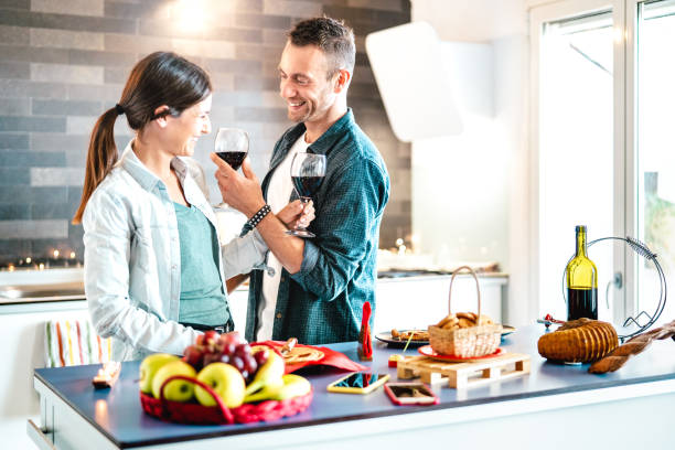 young couple of lovers toasting red wine at house kitchen - happy millenial people enjoying aperitif time cheering together at jubilee anniversary - genuine youth love concept on bright natural filter - wine bottle wine residential structure alcohol imagens e fotografias de stock