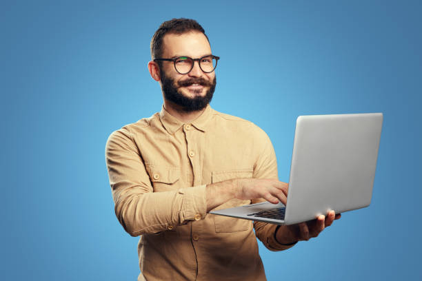 istock Man wearing beige shirt and eyeglasses smiling while holding and using laptop 1347729005