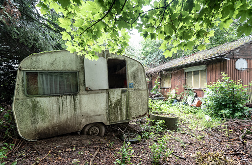 Vintage abandoned caravan amidst greenery against a background of a wooden house and junk around it.
