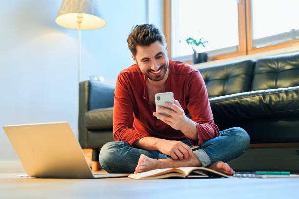 Happy young man using phone while studying at home stock photo