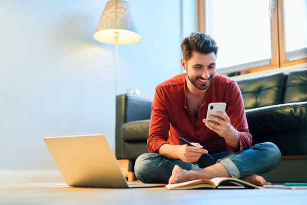 Young man studying at home using smartphone, doing notes stock photo