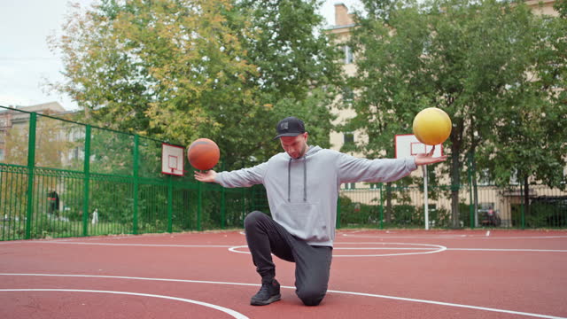 Skilled young man doing basketball freestyle trick spinning two balls on index fingers and kneeling on outdoor college court. Young male freestyler performing stunt with two basketballs