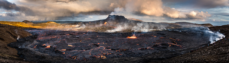 Wide panorama of lava field in front of Fagradalsfjall volcano in Iceland. Crowd of people enjoying the spectacular view in the right side of the picture.