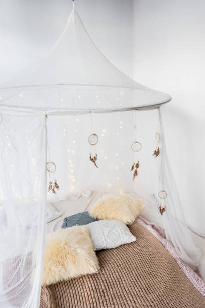 Plants on wooden bedhead of bed with patterned bedding and white drapes in bright bedroom interior with grey bench cama con ropa de cama estampada y cortinas blancas en el interior luminoso del dormitorio. acogedor interior de dormitorio escandinavo en cabaña mansarda. estilo boho, cama decorada a cuadros de lana merino, manta de punto de hilo, guirnalda