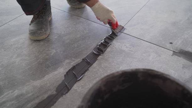 The hand of man holding a rubber float and filling joints with grout. A worker uses black grout to rub the gaps between the tiles on the floor. A worker is rubbing the tiles on the floor stock photo