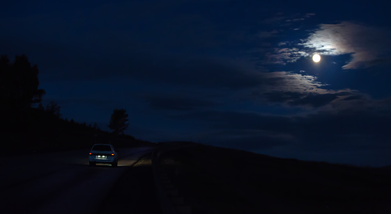 Car moves at night on a road under a cloudy sky lit by the moon