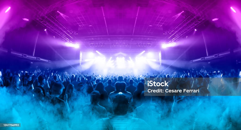 Crowded concert venue Image shot during a music festival. Light comes from a stage with a band show, people silhouettes are visible in front of it. Nightclub Stock Photo