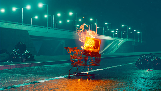 Shopping cart is left in the street and is set on fire. There is an old tv in cart that burns. Garbage bags lays in the street. Freeway is visible in the background.