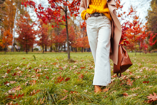 Fashionable woman wearing stylish clothes and accessories walking in fall park. Autumn female outfit. Trendy flared pants with sweater, coat and handbag