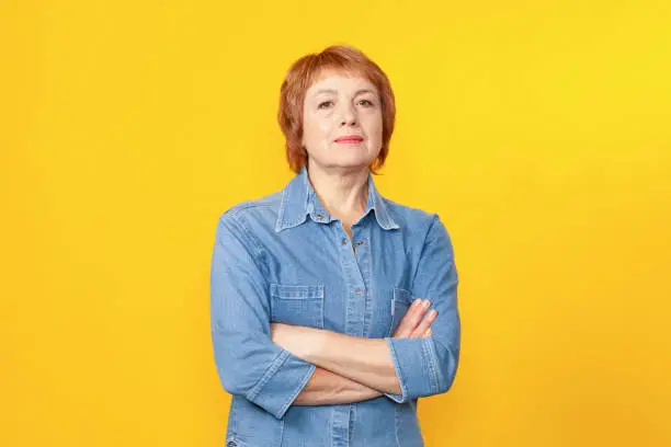 Close up studio portrait of attractive 60 year old woman with short red hair in denim shirt on yellow background