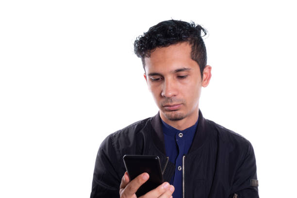Young Latino man, serious and looking at his cell phone isolated on white background. Handsome man reading on his smart phone.