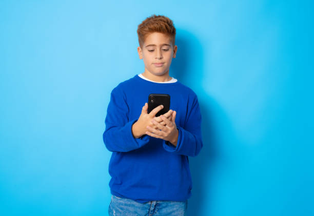 istock Red haired boy in casual sweater using smartphone isolated over blue background. 1347777357