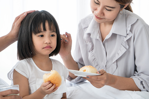Unhappy Asian little girl feeling sick lethargic don't want to eat. young mother take care of her cute daughter and feeds the children breakfast in warm bedroom. happy family On vacation - concept